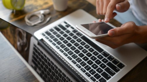 Man Using Smart Phone at Laptop on Desk