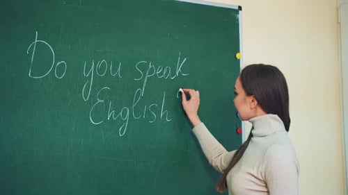 Young teacher writes on a green board with a chalk.