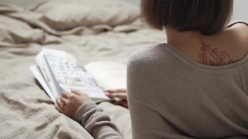 Back View of Slim Lonely Woman with Tattoo Lying in Bed with Book