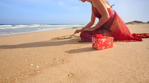 Woman in Red Dress Sits Draws Heart on Sand of Wet Beach