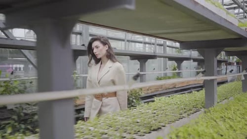 Attractive Young Caucasian Woman Walking in Glasshouse Along Rows of Green Plants and Flowers