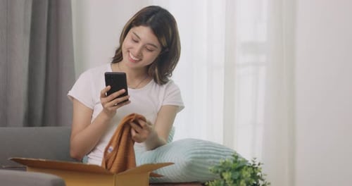 Young Woman Opening Package at Home, Looks at Phone