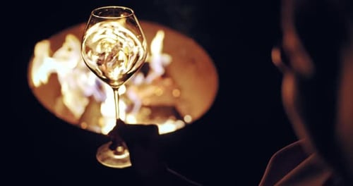 Female Hand Waving Wine in a Glass on Defocused Cozy Camp Fire of Background