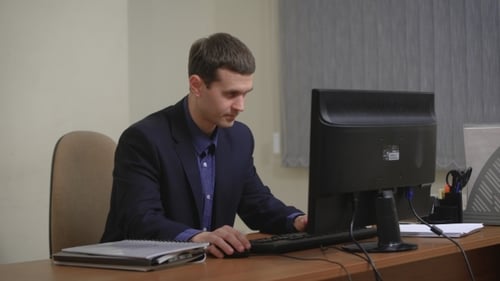 Young Businessman Working In Office, Sitting At Desk, Looking At Computer Screen.