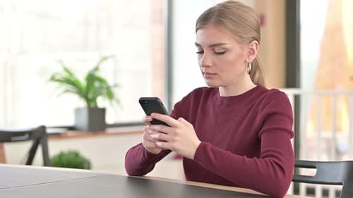 Smartphone Use By Young Woman with Laptop in Office