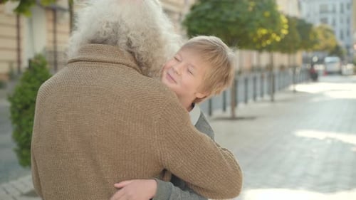 Happy Boy Running To Grey-haired Man Along City Street and Hugging Grandfather. Portrait of Cheerful