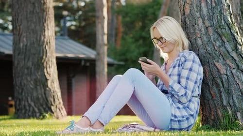 Woman Using Phone Leaning Against Tree on Lawn