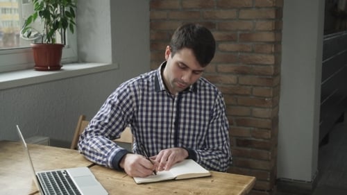 Man Writing in Notebook at Desk with Laptop