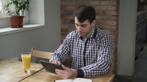 Man Uses Tablet, Drinks Juice at Wooden Table