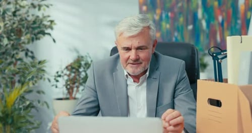 Man in Suit with Gray Hair Leads Remote Conversation From Office Sitting at Desk in Front of Laptop