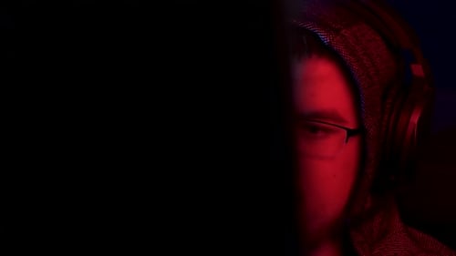 A Young Man Sits at a Computer with Headphones. A Man in a Hood Looks at the Monitor and