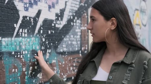 Nachdenkliches junges brünettes Mädchen mit braunen Augen hält ihre Hand an der Wand mit Graffiti und Looks