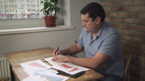 Manager Sitting At Desk, Taking Notes In a Notebook, On a Table Scattered Papers.
