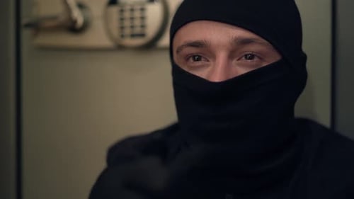 Porträt eines jungen kaukasischen Diebes in schwarzer Kleidung und Sturmhaube, der sich die Hände reibt. Der Typ stiehlt