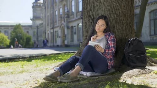 Multinationale junge Frau macht sich Notizen im Notizbuch sitzt unter Baum, Essay