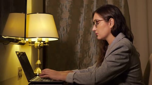 Woman in Glasses and in a Business Suit in the Evening Works at a Laptop