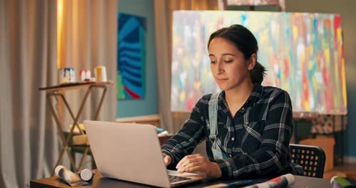 A Smiling Brunette Sits at a Desk in Front of a Laptop in Art Studio