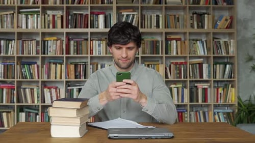 Man Student Using Smartphone Learning Sitting at Books in University Library