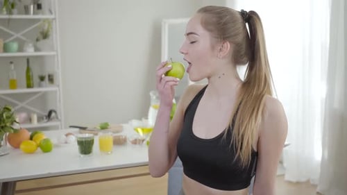 Sportive Young Caucasian Woman Biting Green Apple and Smiling at Camera