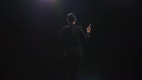 Back View Of Asian Speaker Woman Showing Index Fingers Up While Speaking In The Black Studio