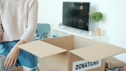 Nahaufnahme weiblicher Hände, die Spendenbox mit Kleidung verpacken, während Frau drinnen zu Hause wohltätige Zwecke tut