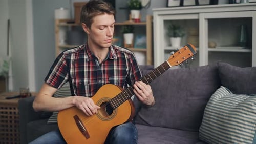 Male Guitarist Is Tuning Acoustic Guitar Checking Sound Touching Strings Sitting on Sofa at Home