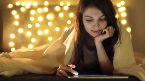 Girl Using Digital Tablet Under Blanket While Lying On Bed At Home