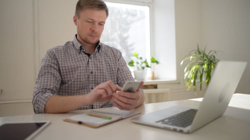 Man Using Smartphone at Desk with Laptop