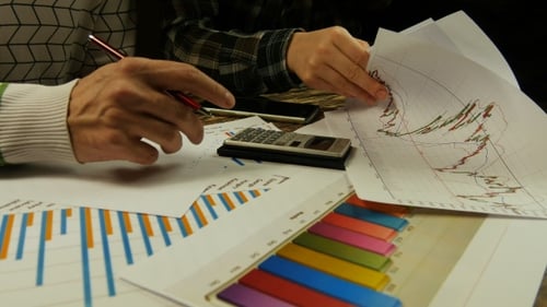 Young Businessman And Businesswoman Analyzing Graphs, Making Calculations And Taking Notes