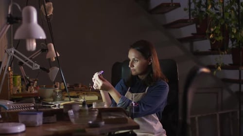 Profile of focused caucasian female jeweller sitting at desk, making jewelry in workshop