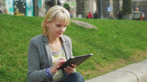 Woman Enjoys The Tablet In The City Sits On a Background Of Green Grass