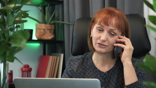 Portrait of confident focused middle-aged businesswoman is talking on phone sitting in modern office