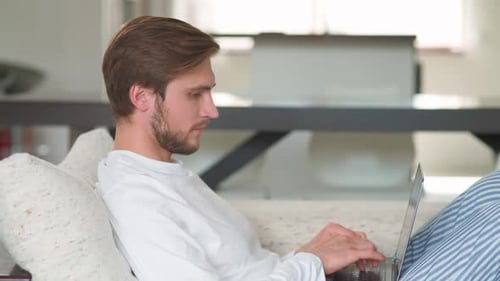 Cheerful Caucasian Man Sits at Home on the Couch and Works Using a Laptop Manager on a Remote Work