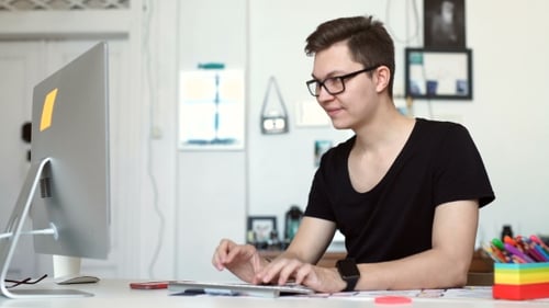 Happy Young Man, Wearing Glasses And Smiling, As He Works On His Laptop