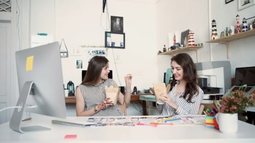 Smiling Young Businesswomen Having Lunch At Table In Office