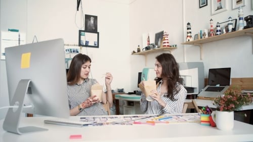 Smiling Young Businesswomen Having Lunch At Table In Office