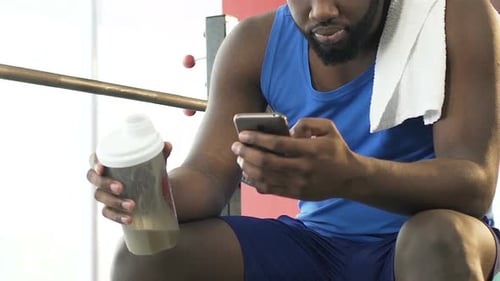 Male Sitting in Gym, Holding Protein Cocktail and Mobile Phone, Social Newsfeed