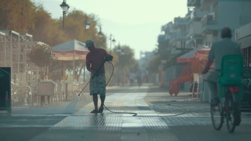 Man Cleaning Asphalt Road Next To Street Cafe With Hose