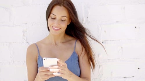 Smiling Woman Using Smartphone in Front of White Wall