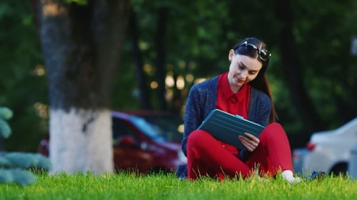 Young Woman Uses Tablet Sitting in Green Park