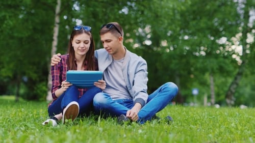 A Young Attractive Couple Sitting On The Grass In The Park, Enjoying The Tablet