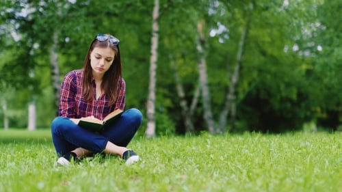 Pretty Young Woman Reading a Book In The Park. He Sits On The Green Grass