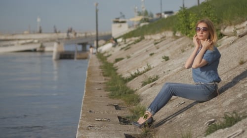 Girl Is Sitting On Quay And Putting On Sunglasses