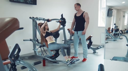 Woman Using Weight Machine with Trainer in Gym