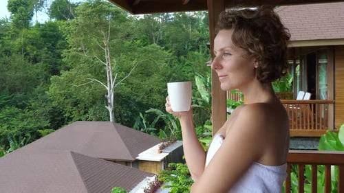 Side View of Beautiful Young Woman Wrapped in a White Bath Towel Stands on Balcony of Villa and