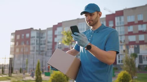 The Postman with Glasses Carries the Parcel and Looks at the Delivery Address Via Mobile Phone