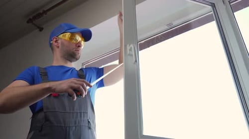 Man in Overalls Measures Width of Window with Tape in House