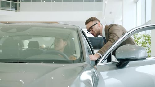 Attractive Girl Talking To Salesman in Motor Dealership Sitting in Modern New Car