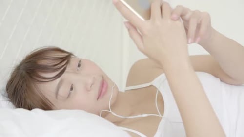 Young Woman Using Smartphone in Bed with Earbuds