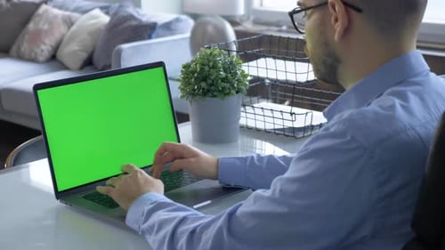 Man Uses Laptop with Green Mock-up Screen While Sitting at the Desk in His Cozy Living Room